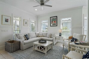 A spacious living room with a white sofa, a coffee table, and a chair. There are framed pictures on the walls, a ceiling fan, and a window with blinds. - Blue Mountain View | Private Pool, Rooftop Deck & Bikes (Santa Rosa Beach)