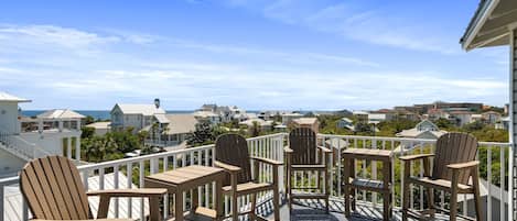 A balcony with wooden chairs and a patterned floor, overlooking a coastal residential area.