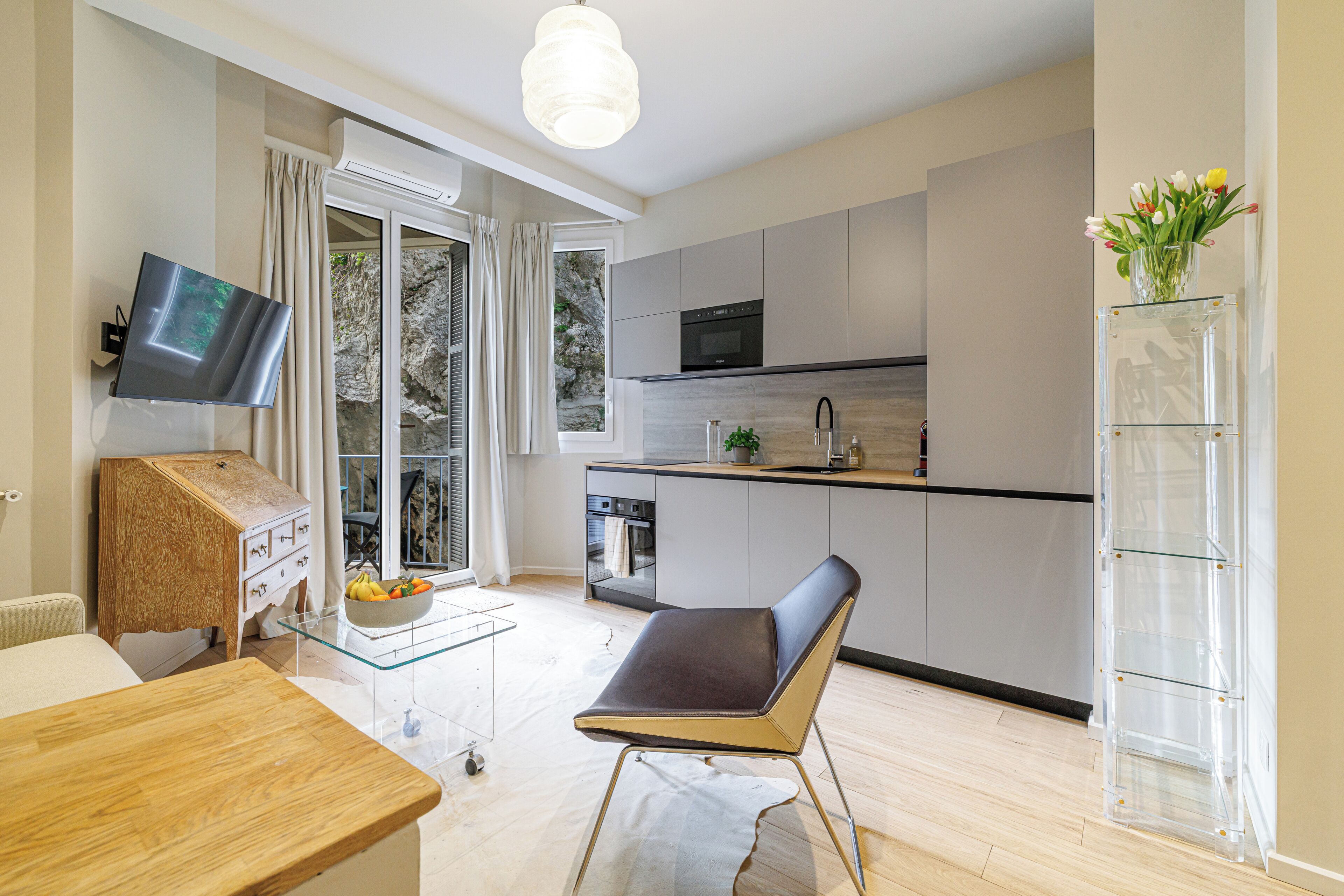 A modern kitchen with a flat-screen TV, a dining area with a table and chairs, and a view of the outdoors through a glass door.