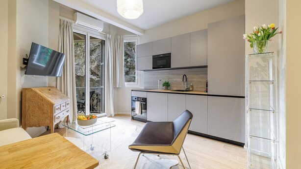 Et moderne køkken med et fladskærms TV, et spiseområde med et bord og stole, og udsigt til det fri gennem en glasdør.