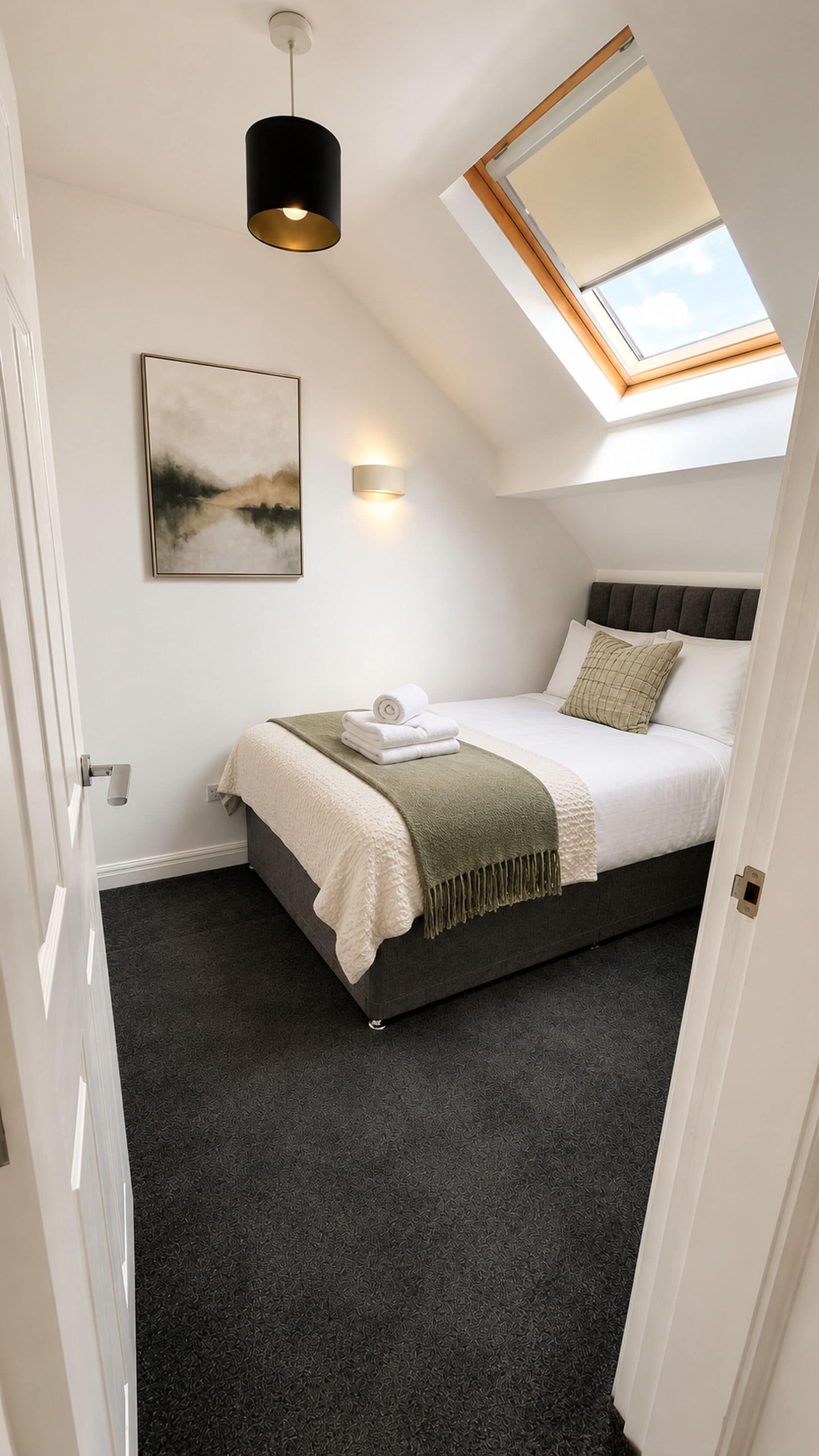 A bedroom with a skylight, a bed with a green throw, a wall-mounted light, and a painting.