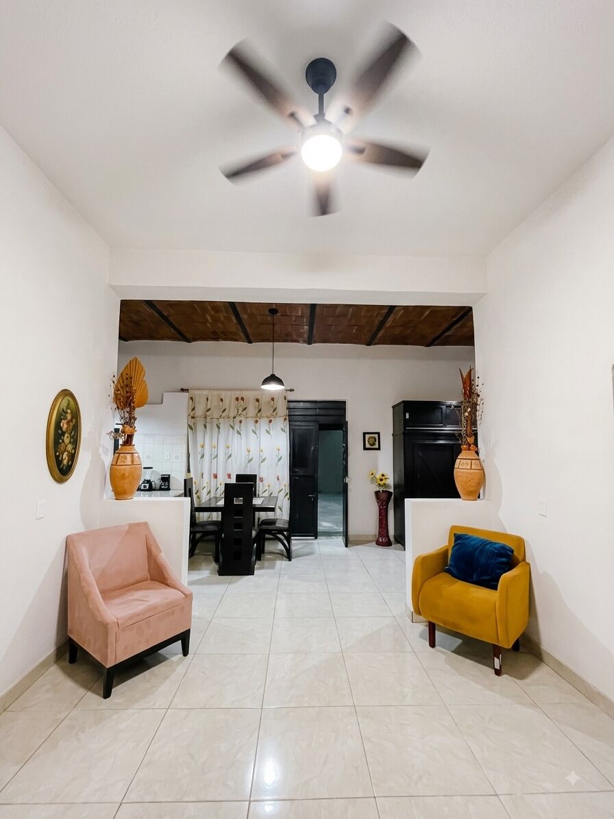 A spacious living area with a ceiling fan, two sofas, a dining table, and a kitchen in the background.