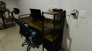 Um pequeno espaço de escritório com uma mesa de madeira, uma cadeira de escritório preta e um computador. Há tomadas elétricas e um filtro de energia na parede.