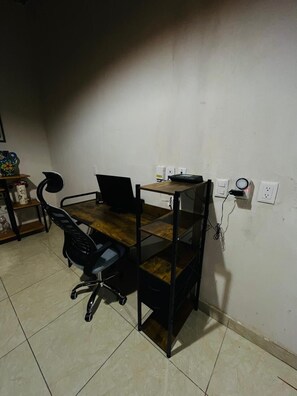 A small office space with a wooden desk, a black office chair, and a computer. There are electrical outlets and a power strip on the wall.
