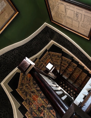 A spiral staircase with ornate carpet and patterned walls, leading to a landing with framed architectural drawings.