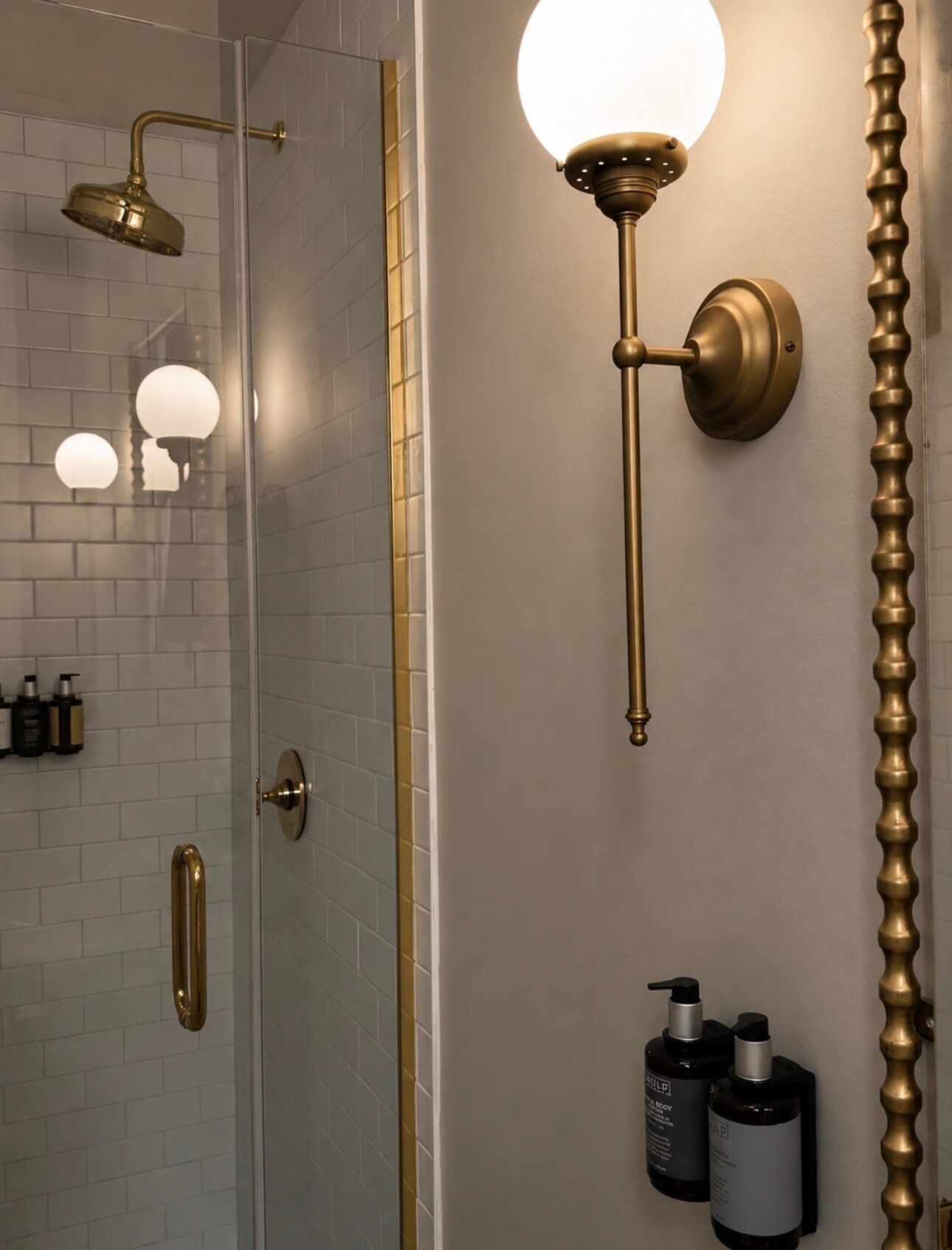 A bathroom with a glass shower enclosure, a wall-mounted light fixture, and toiletries on the wall.