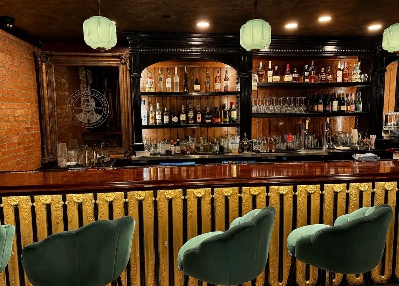 Un espace bar doté d’un comptoir en bois, de tabourets de bar recouverts d’un tissu vert et d’étagères remplies de bouteilles et de verres.
