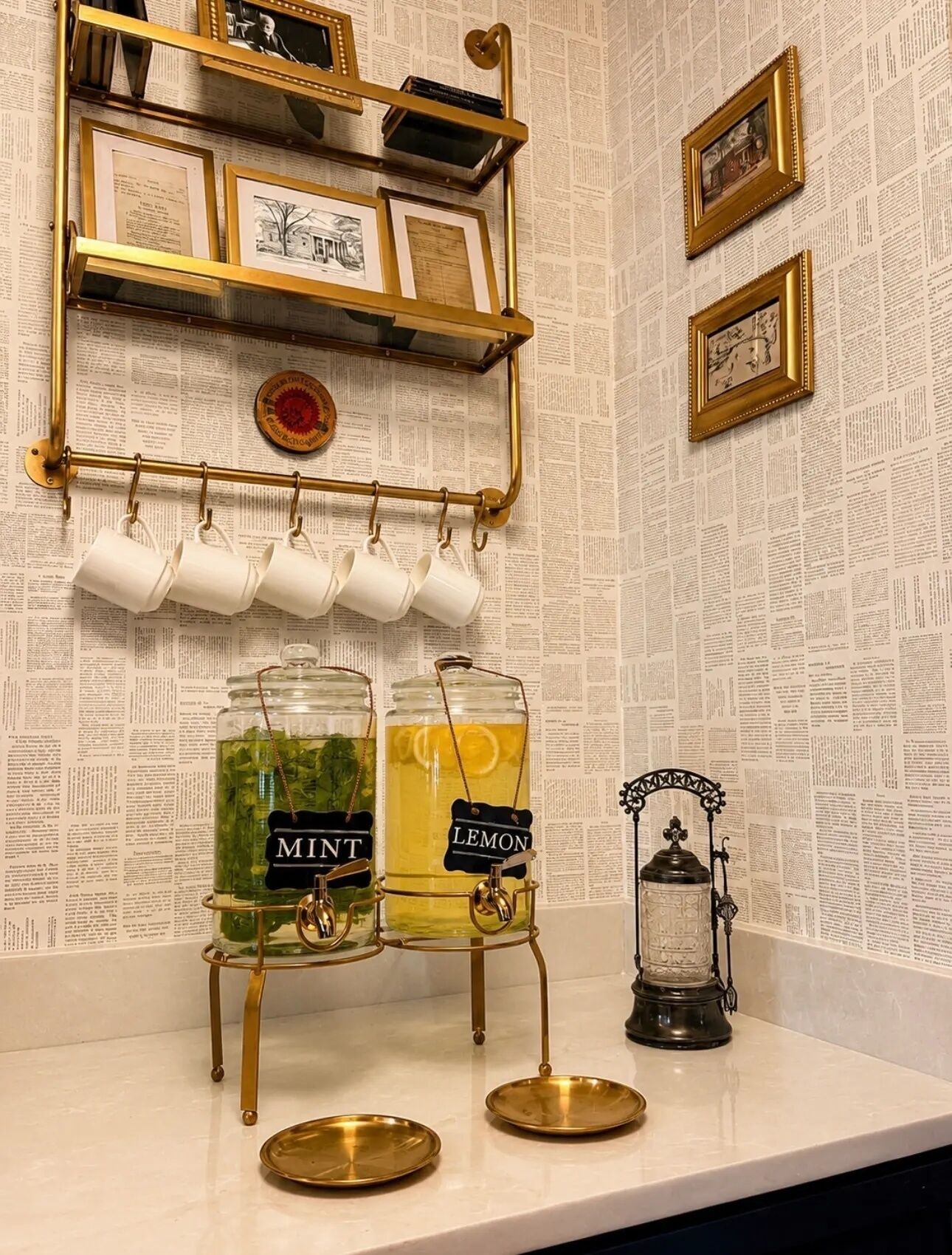 Un taulell de cuina amb dues boles de vidre etiquetades 'MINT' i 'LEMON' que contenen begudes, un suport metàl·lic per a safates amb tasses blanques, una prestatgeria metàl·lica decorativa amb quadres i un fanal d'estil antic.