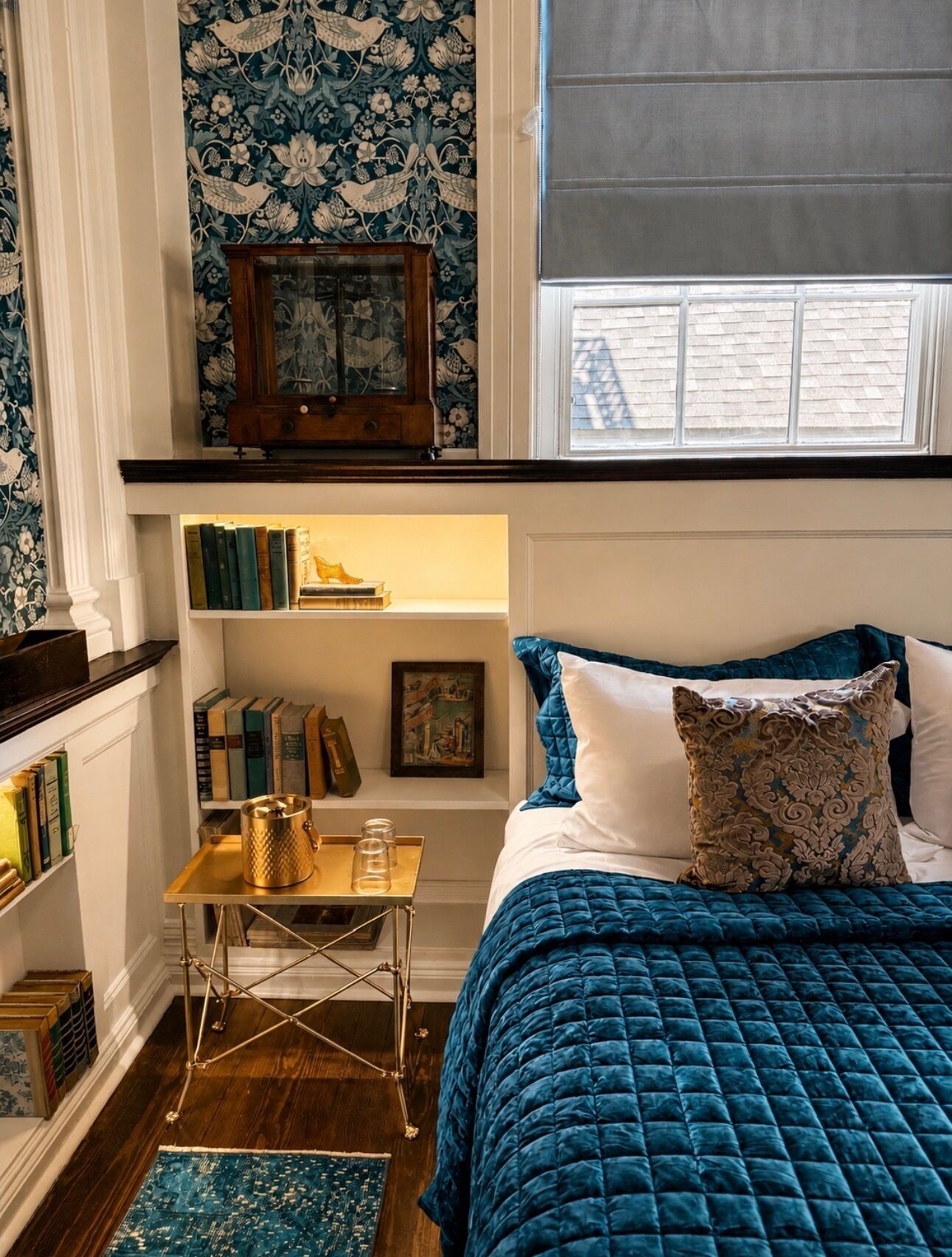 A bedroom with a bed, a small table, a bookshelf, and a window.
