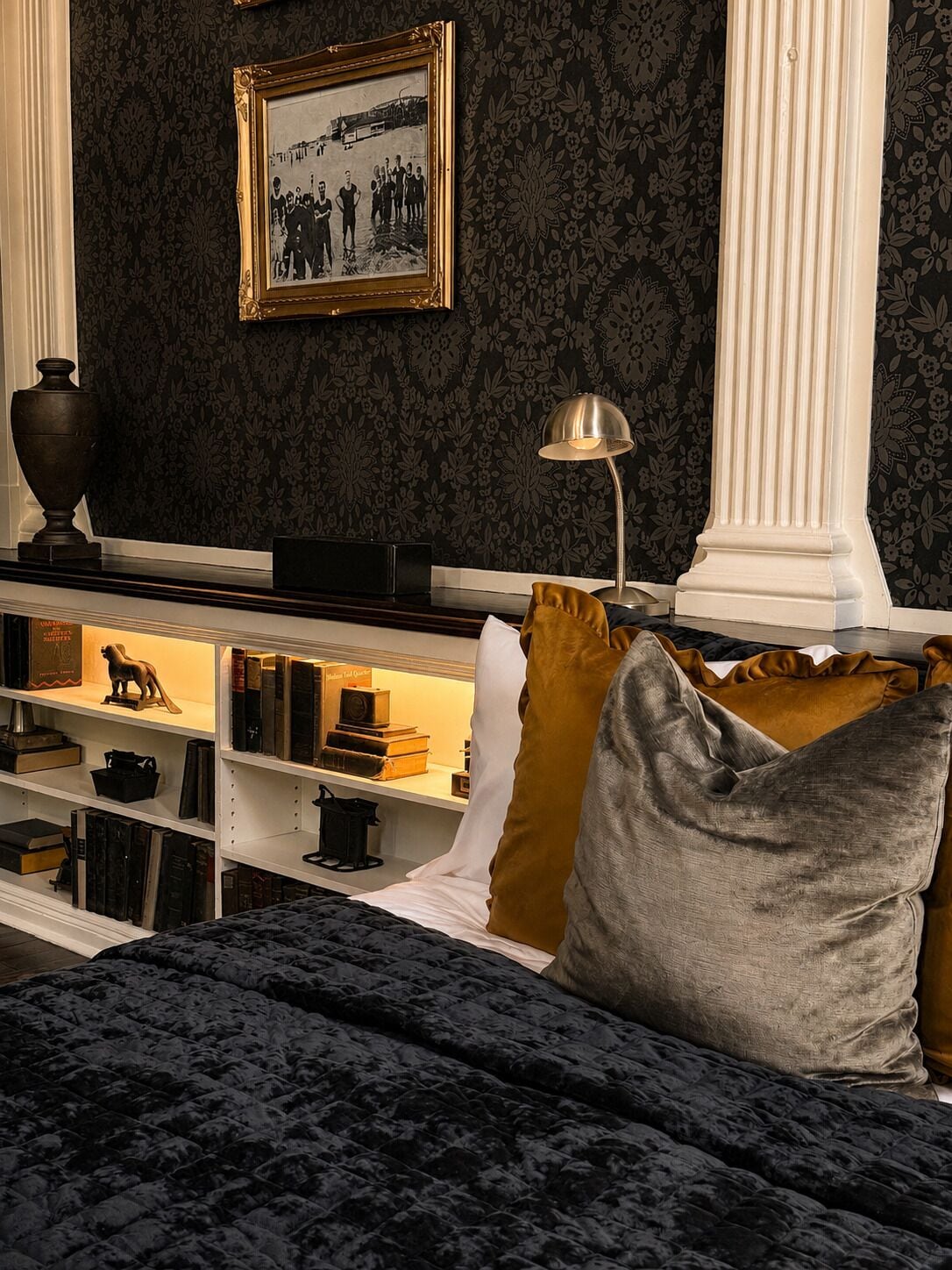 Una habitación con papel tapiz de estampado oscuro, un cuadro en la pared, una estantería con libros y objetos decorativos, una lámpara y una cama con colcha oscura.