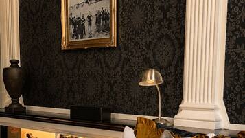 Una habitación con papel tapiz de estampado oscuro, un cuadro en la pared, una estantería con libros y objetos decorativos, una lámpara y una cama con colcha oscura.