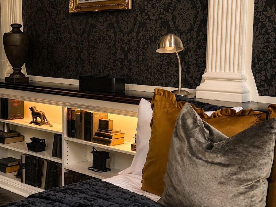 Una habitación con papel tapiz de estampado oscuro, un cuadro en la pared, una estantería con libros y objetos decorativos, una lámpara y una cama con colcha oscura.