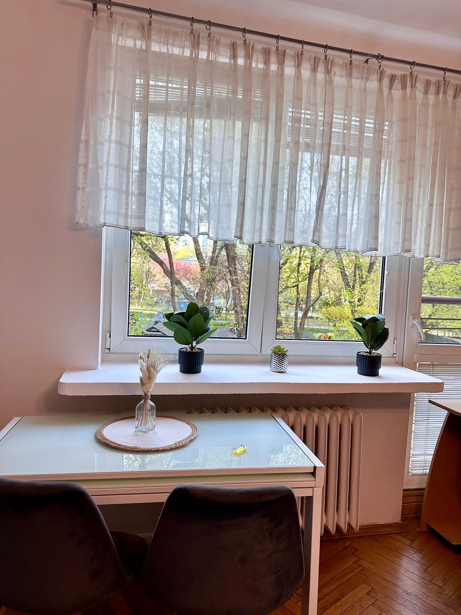 A room with a table and chairs, a window with sheer curtains, and two potted plants on the windowsill.