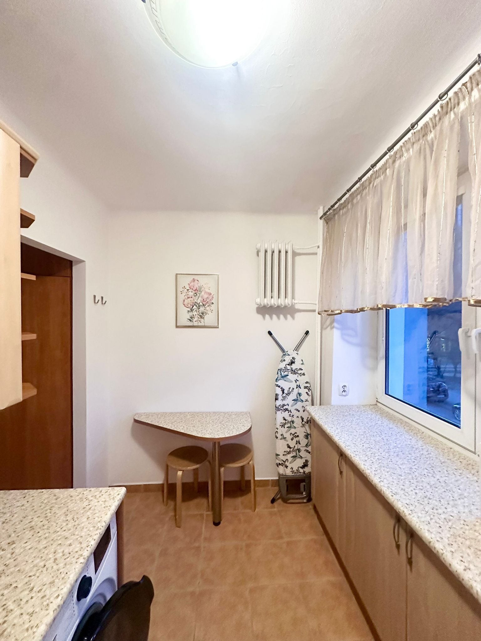 A compact kitchen with a small dining table and stools, a radiator, and a window with curtains.