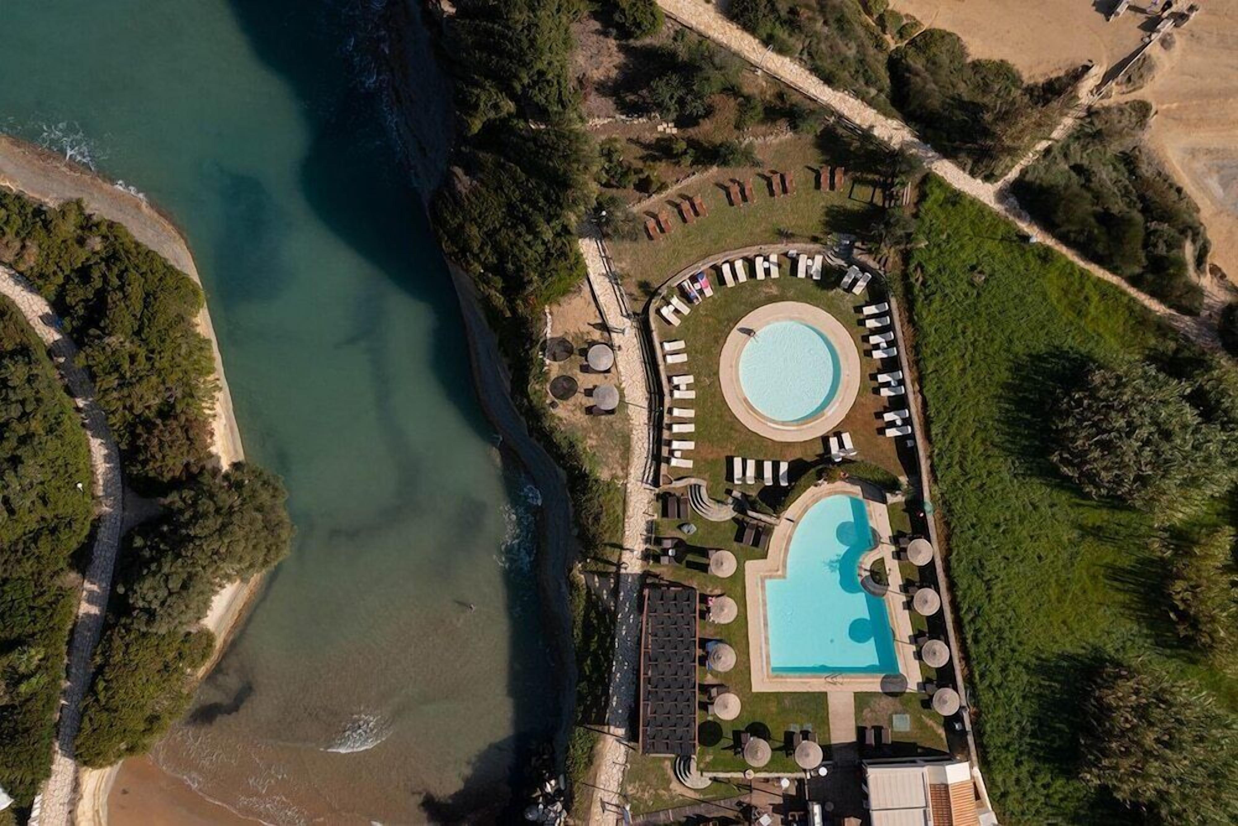 An aerial view of a resort with a central swimming pool, surrounded by lounge areas and a beach.