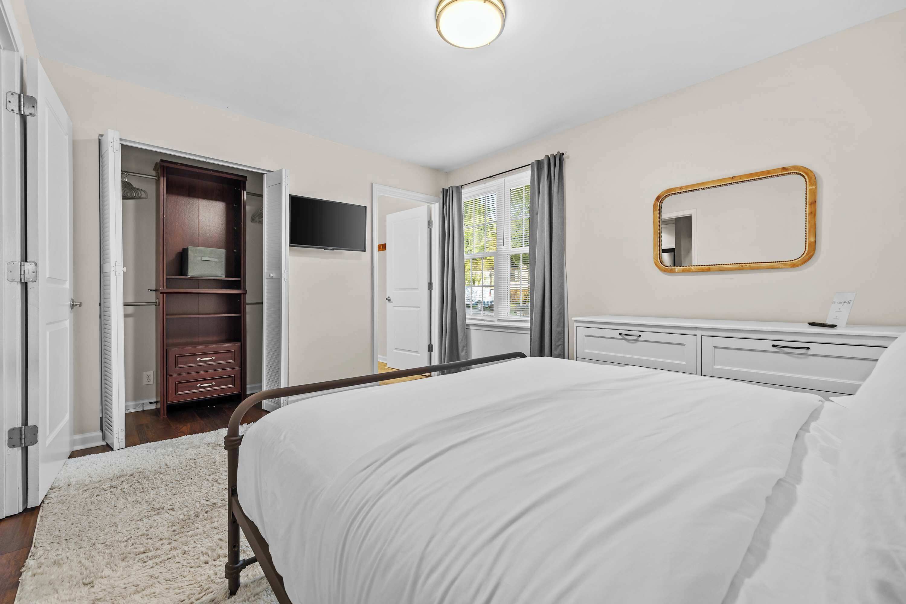 Une chambre à coucher avec un lit, une armoire, une télévision, un miroir et une fenêtre avec des rideaux.
