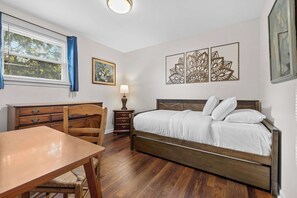 Ein Schlafzimmer mit einem hölzernen Bett, einer Holzkommode, einem Holztisch und einem Fenster mit Jalousien.