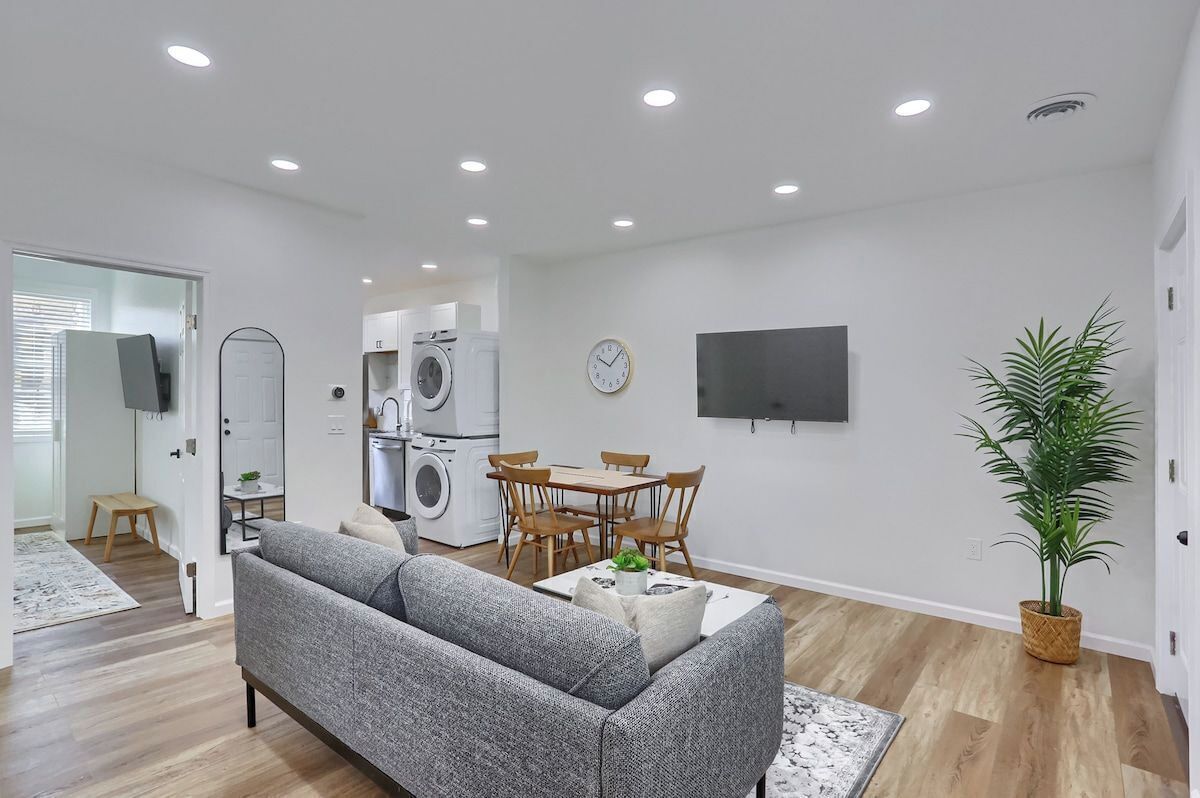 Un salon moderne avec un canapé gris, une table à manger avec des chaises, un téléviseur fixé au mur et un lave-linge à l’arrière-plan.