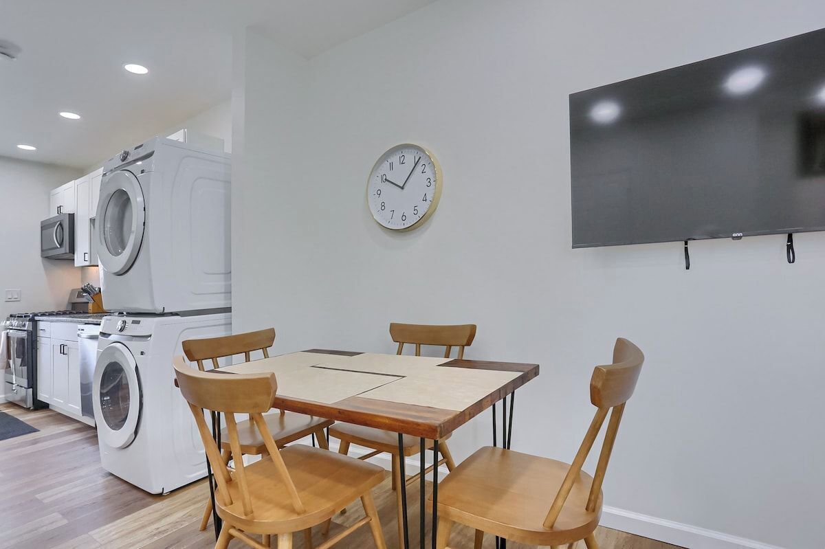 A modern kitchen with a washing machine, a dining table with chairs, a wall clock, and a flat-screen TV.
