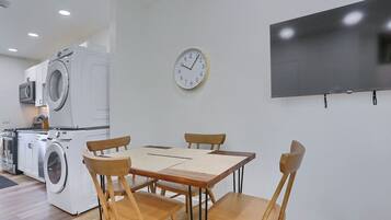 A modern kitchen with a washing machine, a dining table with chairs, a wall clock, and a flat-screen TV.