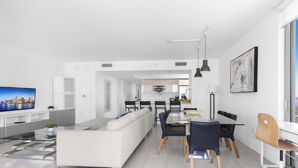 A modern living room with a sofa, dining table, and chairs, a kitchen in the background, and a large window with a city view. - New! 3 BDR Condo Pool Gym - Private Balcony (Hallandale Beach)