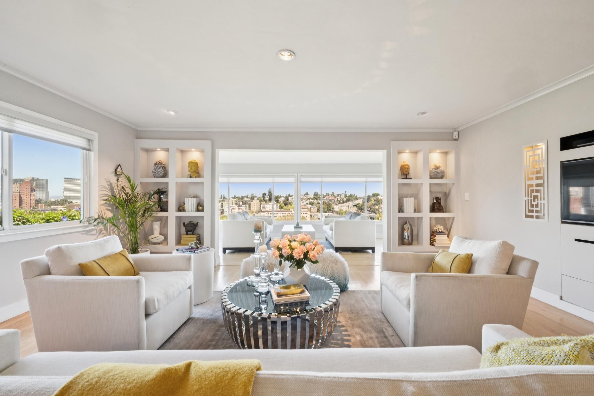 A spacious living room with a city view, white sofas, a round coffee table, and built-in shelving with decorative items.