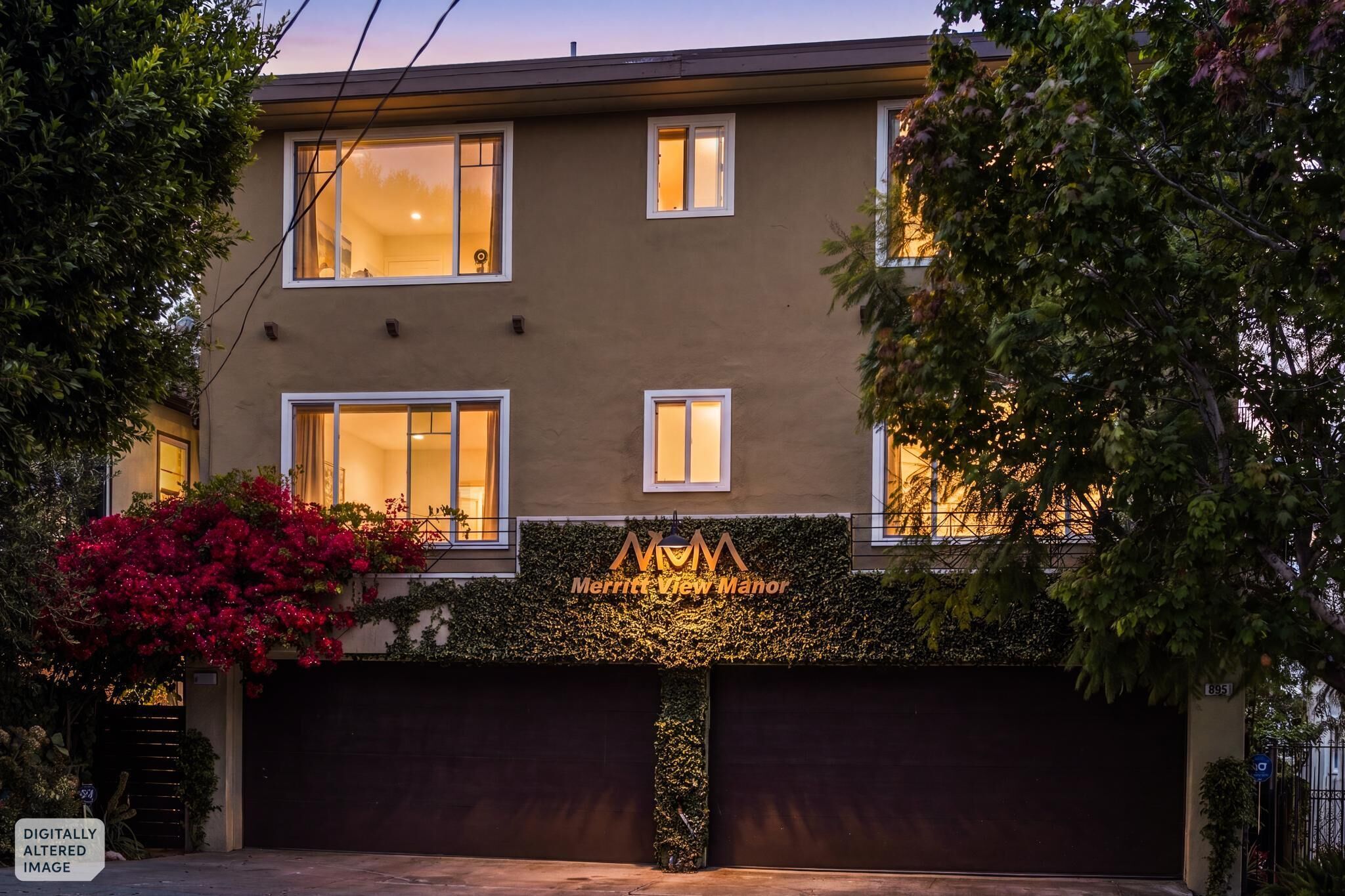 Un bâtiment à deux étages avec une porte de garage de couleur sombre, une enseigne indiquant « Merritt View Manor », et un espace paysager agrémenté de fleurs rouges.