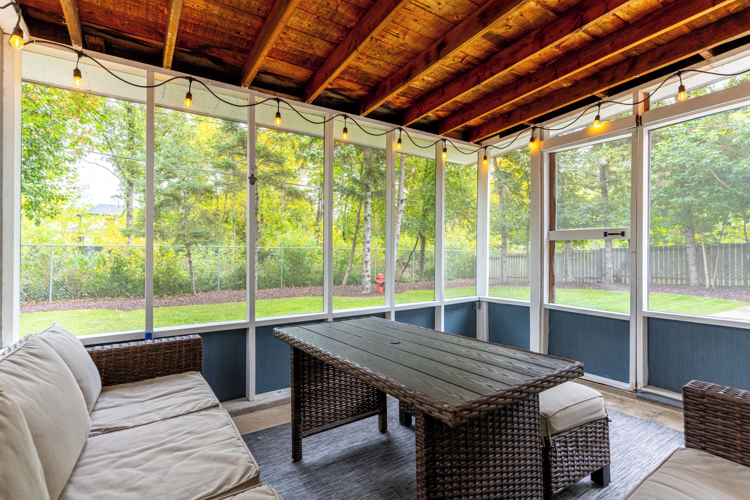 Une terrasse couverte avec une table en bois et des meubles en osier, de grandes fenêtres et une vue sur un paysage verdoyant.
