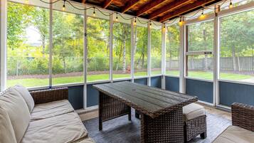 Une terrasse couverte avec une table en bois et des meubles en osier, de grandes fenêtres et une vue sur un paysage verdoyant.