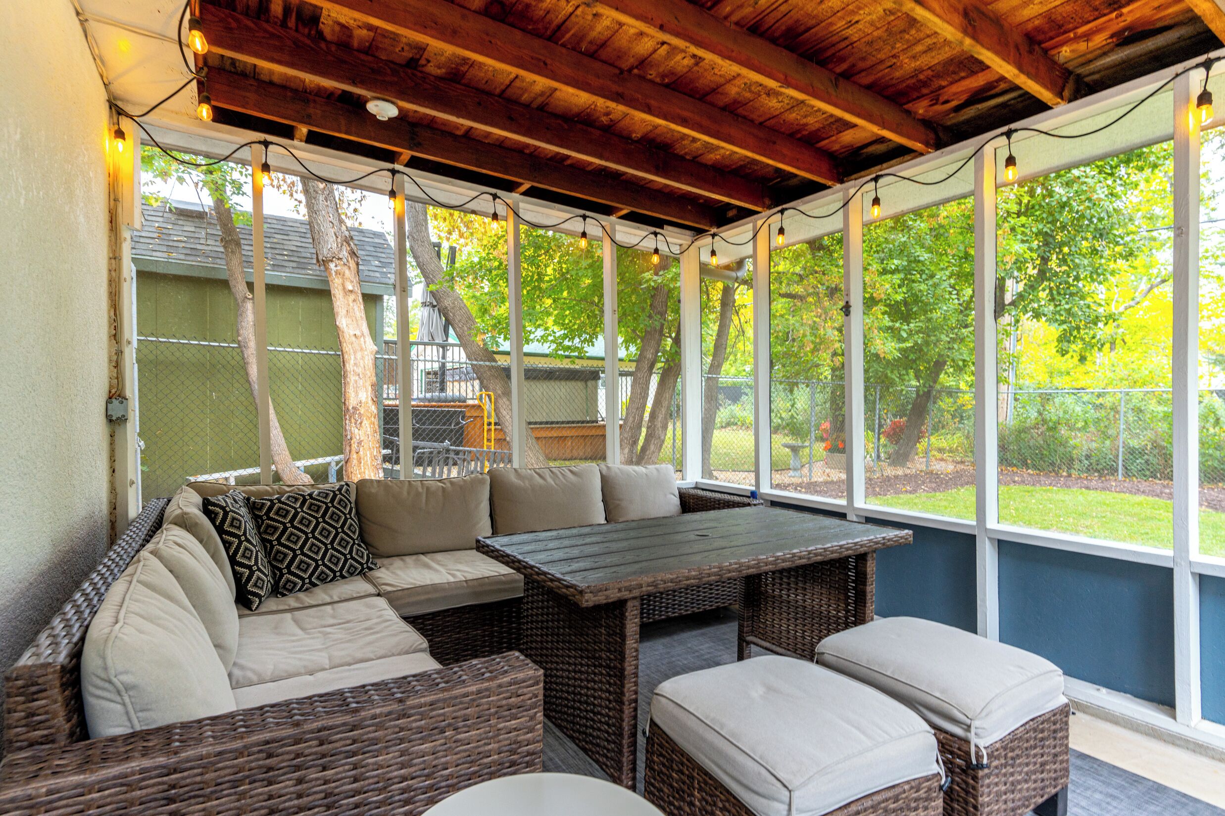 Eine überdachte Terrasse mit Korbmöbeln, einem Tisch und Blick auf einen umzäunten Garten.
