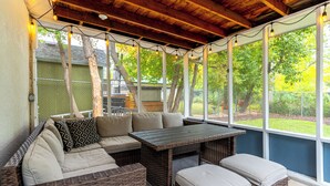Un portico riparato con mobili in vimini, un tavolo e vista su un giardino recintato.