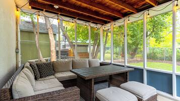 Eine überdachte Terrasse mit Korbmöbeln, einem Tisch und Blick auf einen umzäunten Garten.