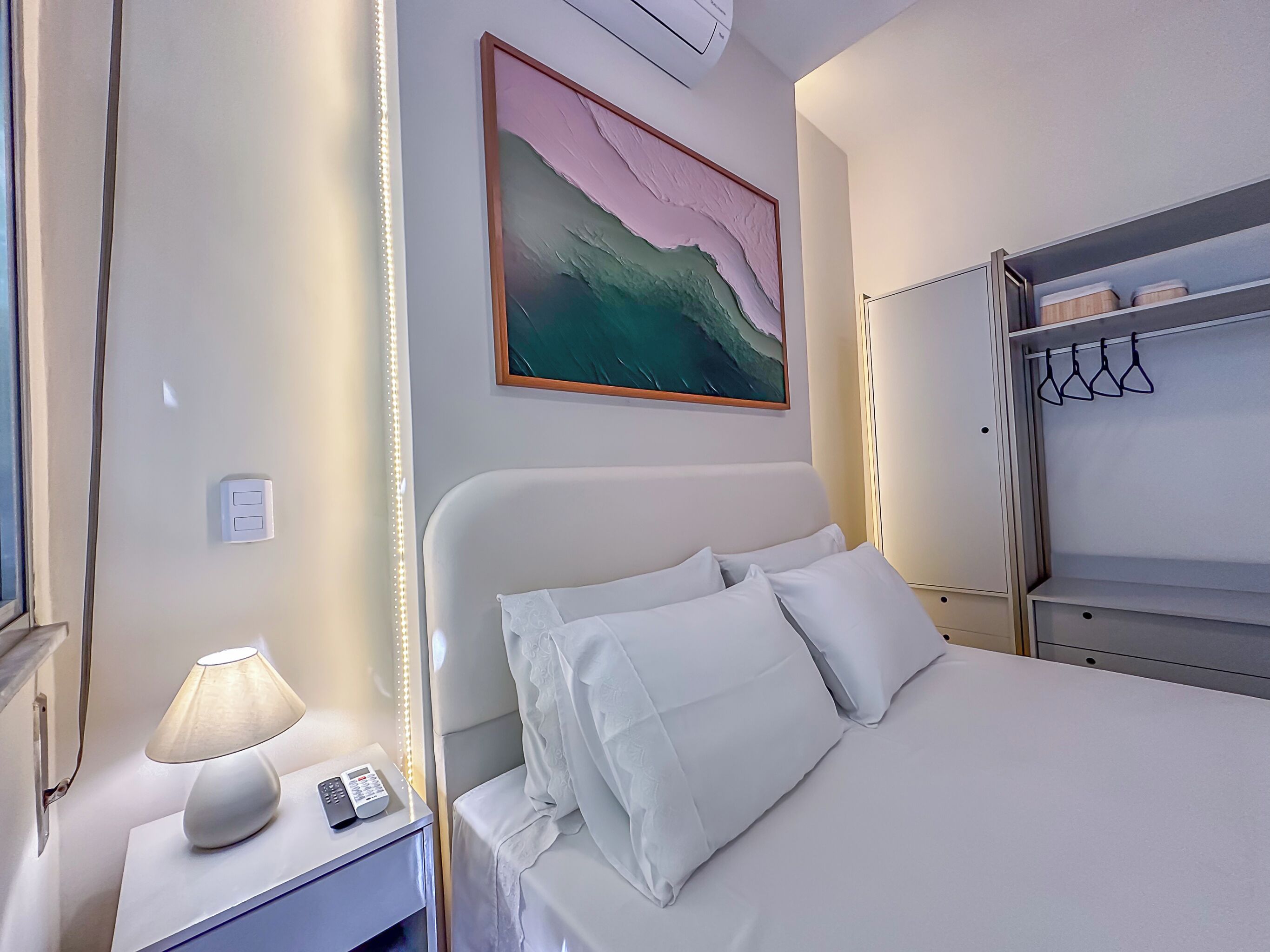 Ein Hotelzimmer mit Bett, Nachttisch mit Lampe und Telefon, einem Bild an der Wand und einem Schrank mit Kleiderhaken.