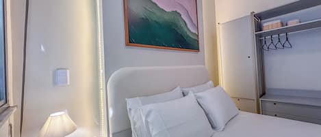 A hotel room with a bed, a bedside table with a lamp and phone, a painting on the wall, and a wardrobe with hangers.