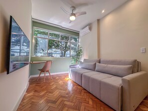 A room with a herringbone wooden floor, a grey sofa, a wooden chair, a ceiling fan, and a window with a view of trees and buildings. - Copacabana: renovated, 3 minutes from the beach (Rio de Janeiro)