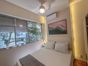 A bedroom with a bed, a window with blinds, an air conditioning unit, a ceiling fan, and a wall-mounted painting. - Copacabana: renovated, 3 minutes from the beach (Rio de Janeiro)