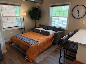 A compact living space with a bed, a sofa, a dining area, and a large window with blinds.