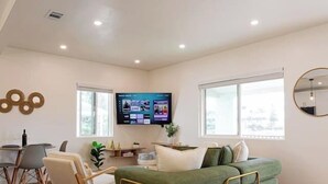 Un salón amplio con un sofá verde, una mesa de comedor con sillas, un televisor de pantalla plana montado en la pared y grandes ventanas que dejan entrar luz natural.