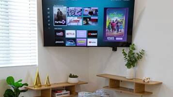 Een woonkamer met een flatscreen-tv aan de muur, waarop diverse streamingdiensten te zien zijn. Er staat een rond rieten bijzettafeltje met een schaal lekkernijen, een bank en een bloempot met plant.