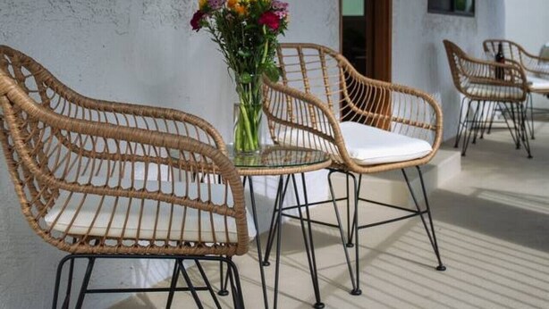 Sillas de mimbre con cojines, una mesa de vidrio y un jarrón con flores en un balcón con vistas al cielo.