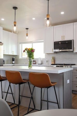 A modern kitchen with white cabinets, a central island with stools, a built-in microwave, and pendant lighting. - Luxury 2BR Escape - Steps to Beach & Restaurants (Oceanside)