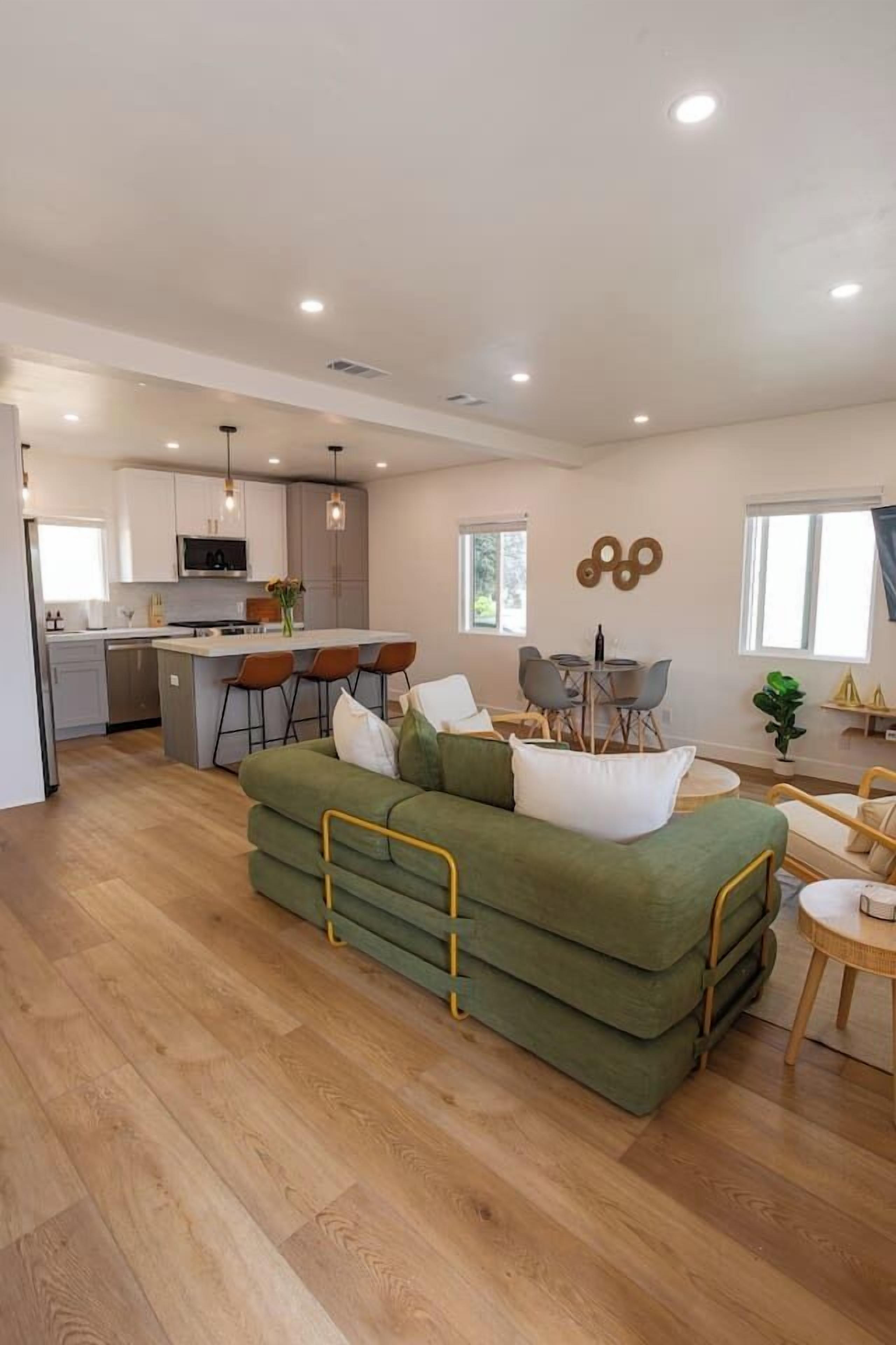 A modern living room with a green sofa, a dining area with a table and chairs, and a kitchen in the background.