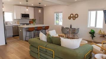 A modern living room with a green sofa, a dining area with a table and chairs, and a kitchen in the background.