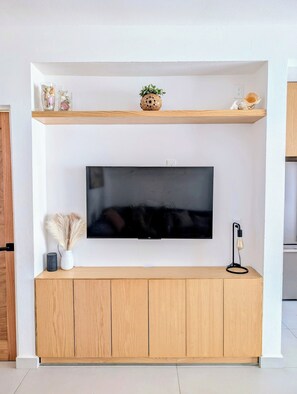 A flat-screen TV mounted on a wall above a wooden cabinet with a small plant and decorative items on the shelf above. - Modern Coastal Oasis with Private Pool (Puerto Morelos)