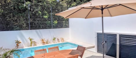 Un espace aménagé au bord de la piscine, avec une table et des chaises, un luminaire suspendu et un grand parasol.