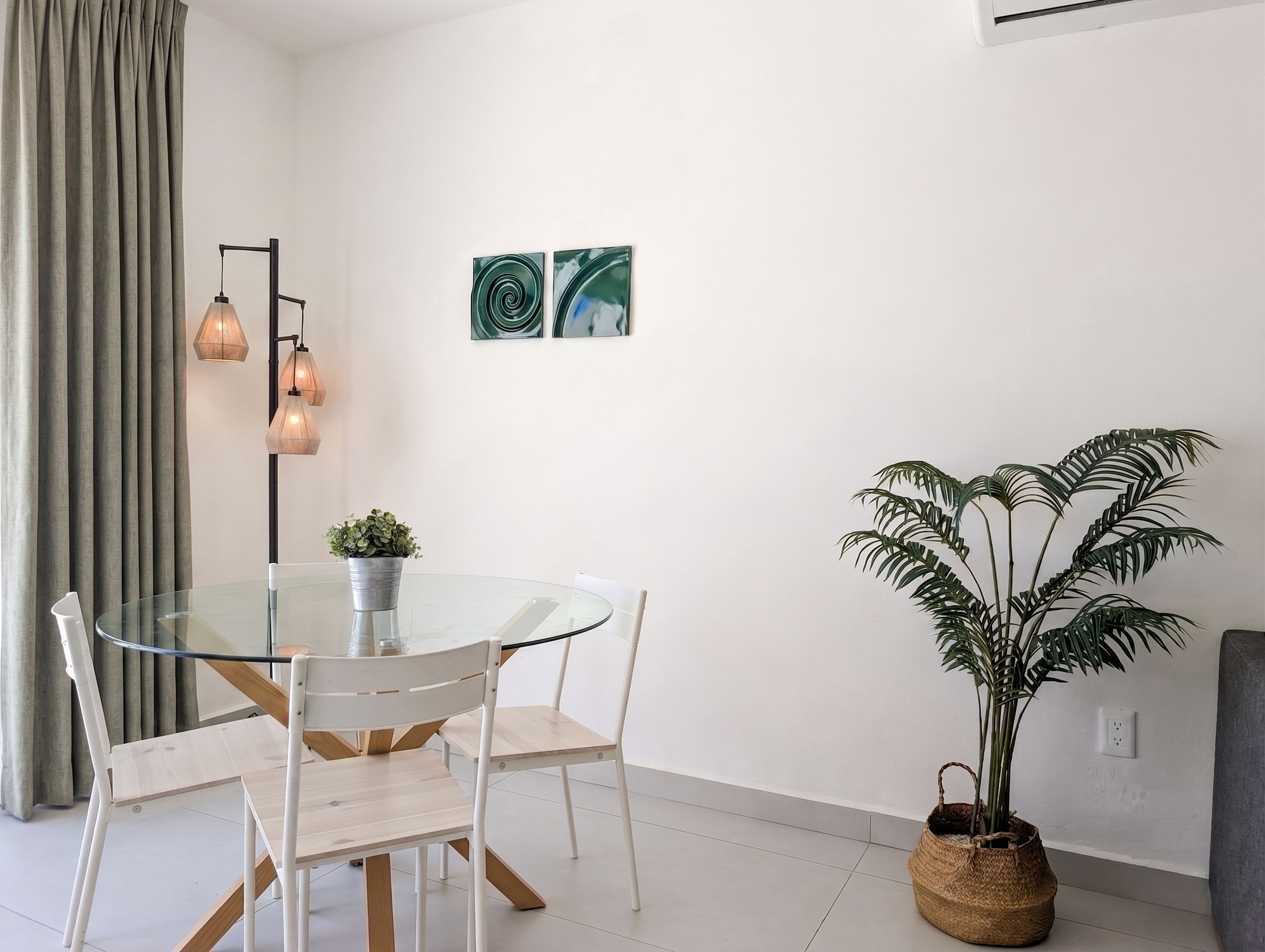 A modern dining area with a glass table, white chairs, a potted plant, and two wall art pieces.