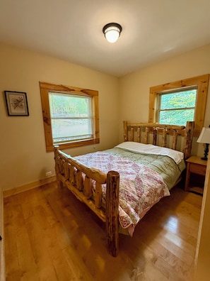 A bedroom with a wooden bed frame, a quilted bedspread, two windows with blinds, a bedside table with a lamp, and a ceiling light fixture.