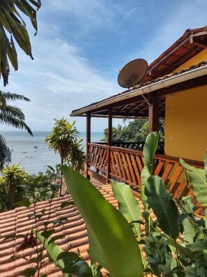 A terrace with a view of the sea, surrounded by tropical plants and a wooden structure with a satellite dish.