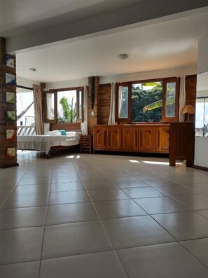 A spacious room with a bed, a wooden cabinet, and large windows offering a view of greenery.