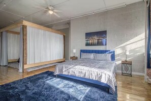 Ein Schlafzimmer mit einem Bett, einem Deckenventilator, einem Nachttisch, einer Lampe, einem blauen Teppich und einem Stadtbild an der Wand.